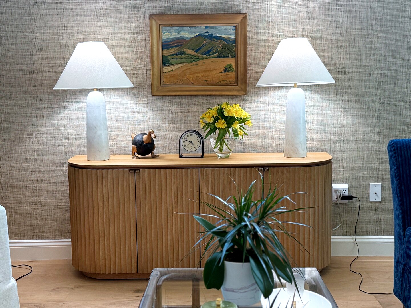 Sideboard vignette with a framed landscape painting, marble lamps, a bronze bird, a clock, and yellow flowers