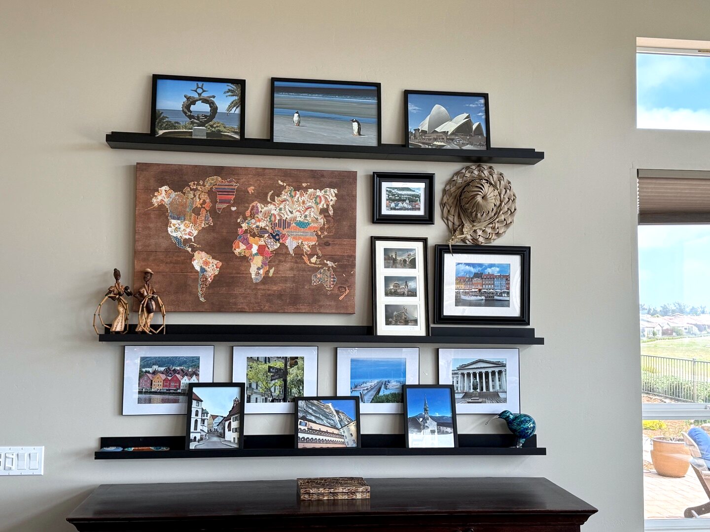 Completed travel gallery wall — three floating shelves with a fabric world map, figurines, and framed travel photographs
