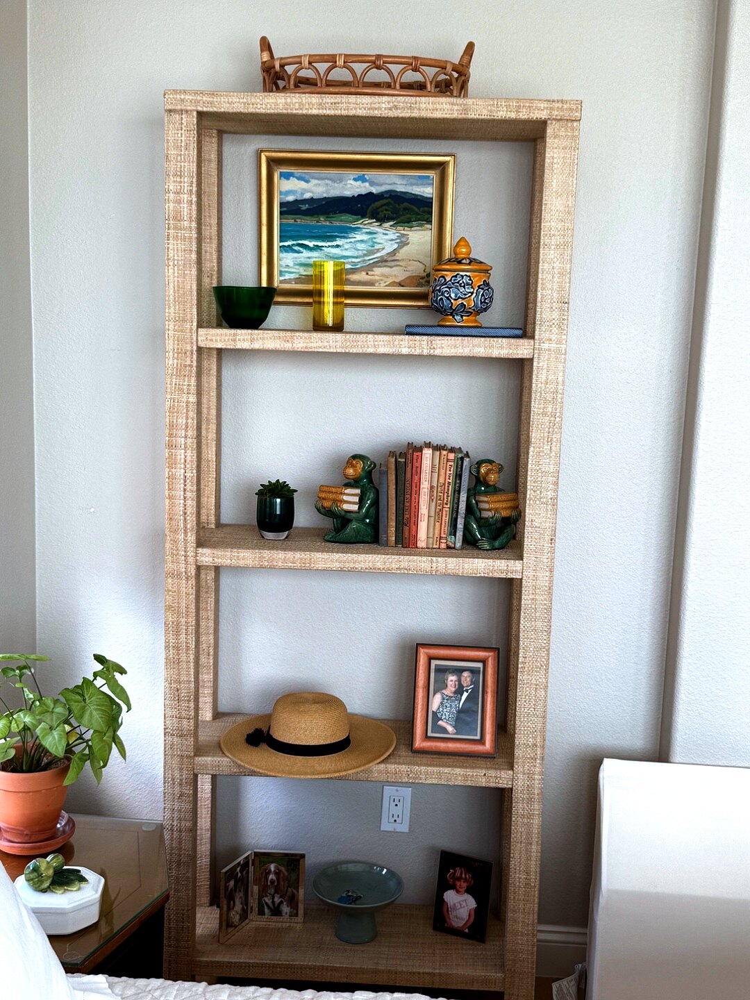 Raffia etagere styled with a coastal painting, ginger jar, monkey bookend, and family photographs